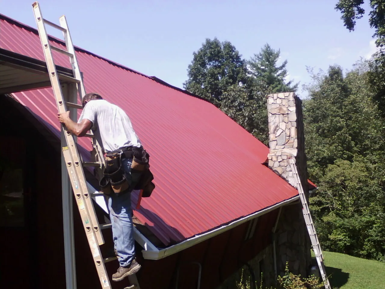 Roofer on a ladder beside a red metal roof and stone chimney — Soffit & Fascia Repair by Roofing Brothers in Hilliards